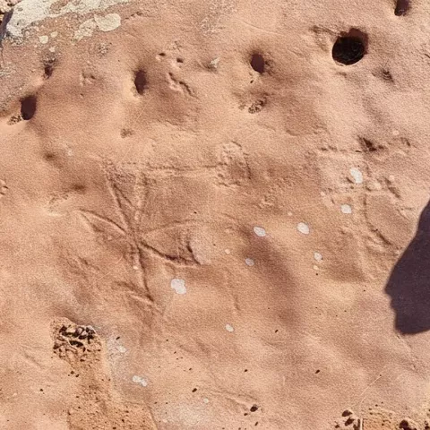 Grabados rupestres en roca rojiza con figuras geométricas y pequeños orificios.