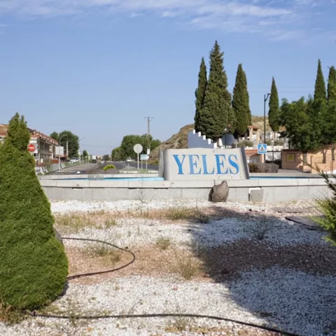 Rotonda de entrada a un pueblo con un cartel de letras azules, rodeada de arbustos decorativos, piedras y cipreses bajo un cielo despejado.