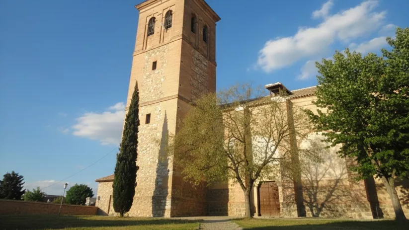 Una toma de ángulo bajo muestra la alta torre de ladrillo y piedra de una iglesia con una aguja cónica y un campanario de dos niveles, rodeada por árboles verdes en un día soleado.