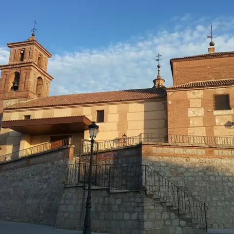Vista exterior de una iglesia de ladrillo con un campanario a la izquierda, escaleras de piedra que suben a la entrada principal y un gran muro de contención de piedra.