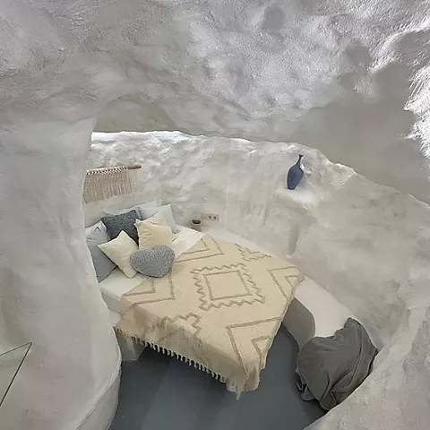 Dormitorio excavado en roca con cama, textiles claros y chimenea encendida.