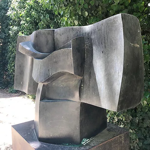 Escultura abstracta de metal oscuro con formas curvas sobre pedestal al aire libre.