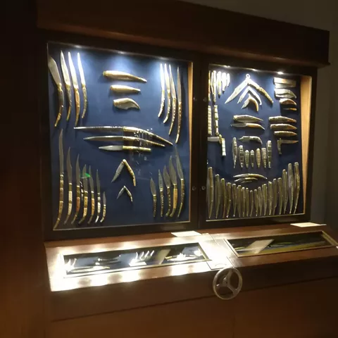 Vitrina de museo con navajas y cuchillos antiguos iluminados y organizados por tipos.