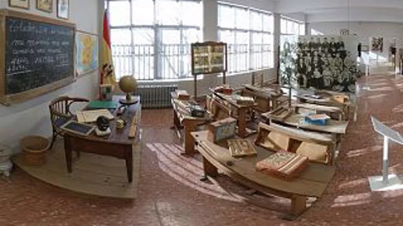 Vista panorámica de aula antigua con mesas, mapas, libros y pizarras.