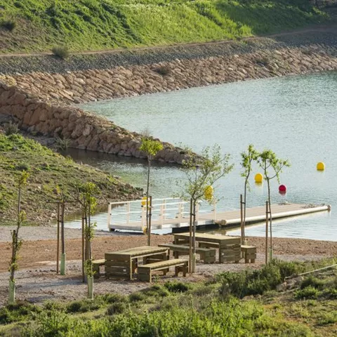 Zona recreativa junto al agua con pasarela, bancos y vegetación.