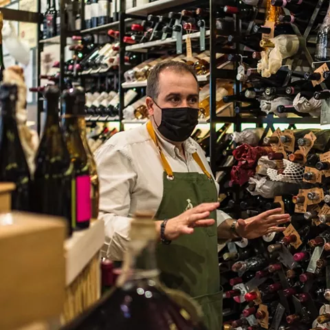 Hombre con delantal y mascarilla gesticula entre estanterías repletas de botellas de vino.