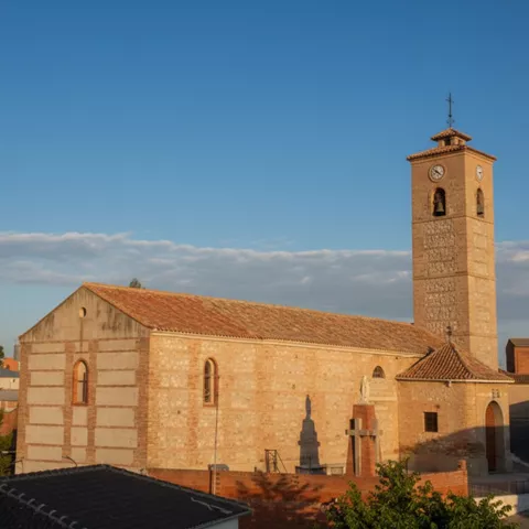 Una iglesia rural de piedra y ladrillo con un tejado de tejas rojas y un campanario alto con un reloj, bajo un cielo azul claro con nubes suaves.