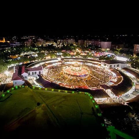 Vista aérea nocturna de un recinto circular iluminado, con gran afluencia de personas y luces festivas.