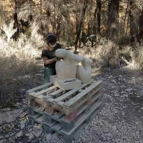Persona trabajando una escultura de piedra al aire libre entre árboles.