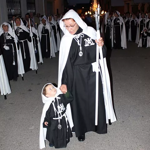 Mujer y niño con túnicas oscuras y cirios encabezando una procesión nocturna.