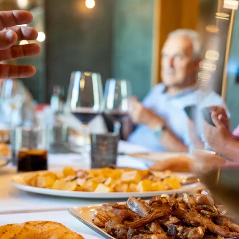 Mesa con platos y pan en primer plano, comensales desenfocados al fondo.