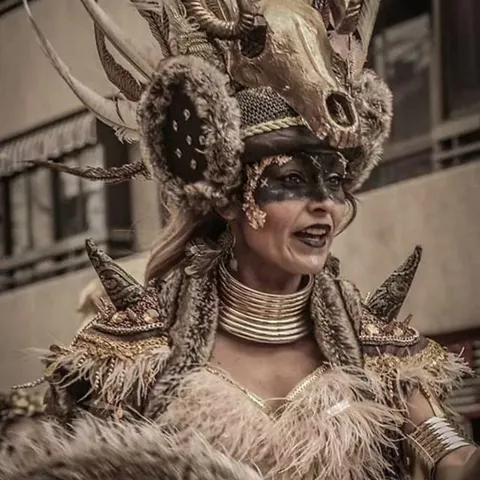 Persona con maquillaje oscuro y tocado con cuernos y plumas en un desfile.
