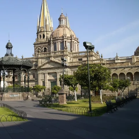 Jardín urbano con quiosco ornamentado y catedral al fondo.
