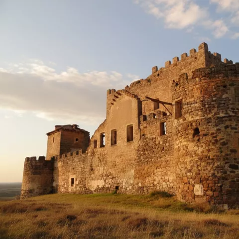 Vista lateral de murallas de piedra con torre redonda y ventanas, bajo cielo con nubes.