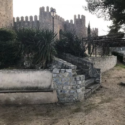 Muralla almenada vista desde un jardín con escalinata y pérgola.