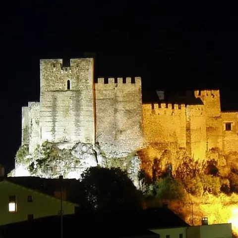 Castillo amurallado iluminado de noche sobre un cerro rocoso.