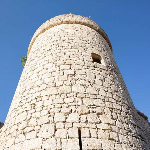 Torre circular de piedra vista desde abajo, con muro defensivo y cielo azul de fondo.