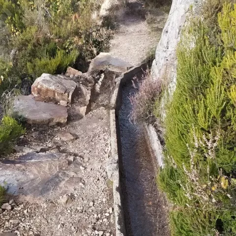 Sendero junto a canal de agua entre rocas y matorral.