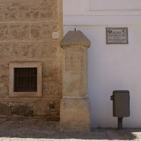 Fachada de piedra y ladrillo con ventana enrejada y columna con inscripción junto a muro blanco.