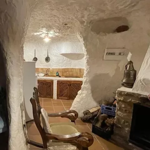 Interior de vivienda excavada en la roca con chimenea, sillón antiguo y cocina al fondo.