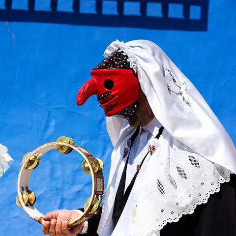 Persona enmascarada tocando una pandereta junto a flores decorativas.