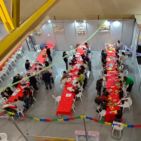 Vista cenital de un taller infantil con mesas largas, manualidades y muchas personas sentadas.