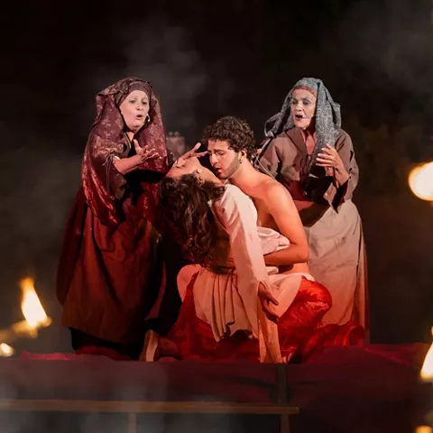 Escena dramática con varios actores en vestuario tradicional rodeados de velas y ambiente oscuro.