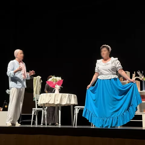 Dos personas en escena junto a una mesa con flores, con una mujer levantando la falda azul.
