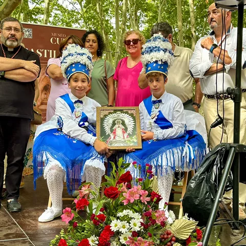 Dos niños con trajes tradicionales sostienen un cuadro religioso en un escenario al aire libre.