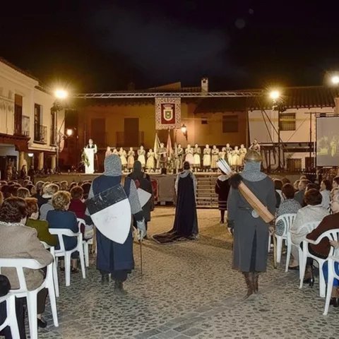 Acto nocturno al aire libre con escenario iluminado, público sentado y figuras con armadura en primer plano.