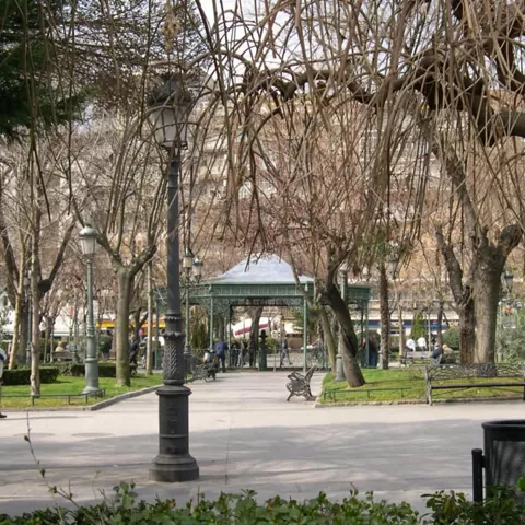 Parque urbano con paseo central, arbolado y quiosco metálico.
