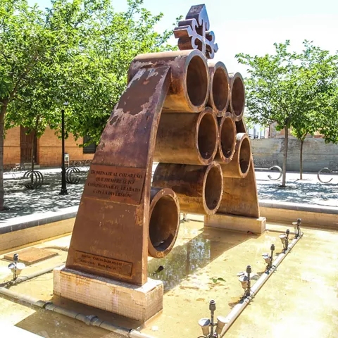 Escultura metálica sobre una fuente en una plaza arbolada.