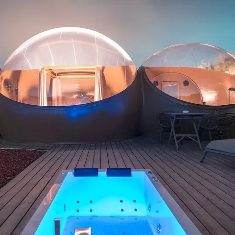 Alojamiento de cúpulas transparentes con jacuzzi iluminado en una terraza al anochecer.