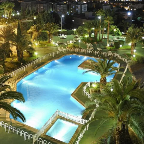 Piscina exterior iluminada de noche con jardines y vistas urbanas.