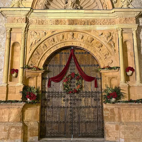 Portada de piedra tallada con relieves y gran puerta de madera decorada con corona floral.