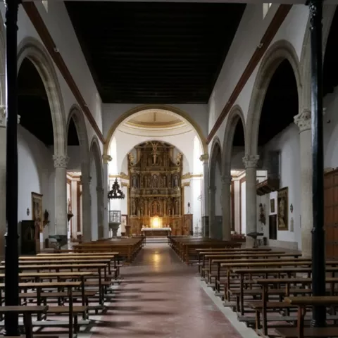 Interior de iglesia con largas hileras de bancos y retablo al fondo.