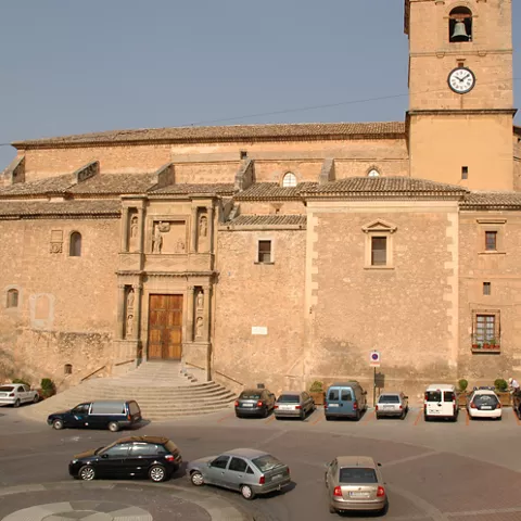 Vista frontal de iglesia de mampostería con torre y reloj, con coches aparcados delante.