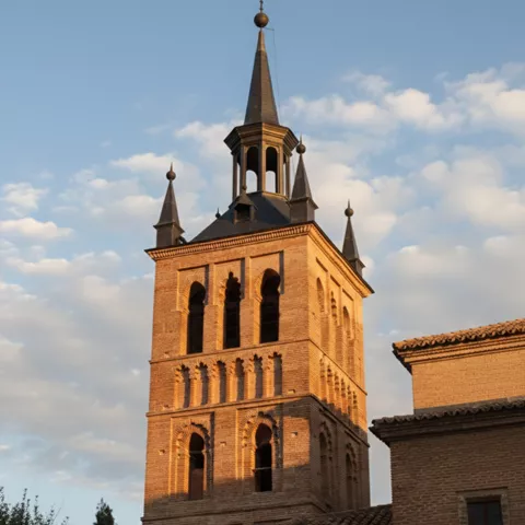 Torre campanario de ladrillo con arcos y chapiteles iluminada al atardecer.