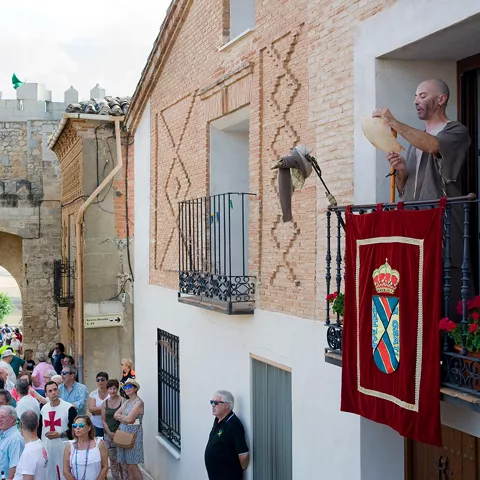 Persona en balcón tocando instrumento ante puerta amurallada.