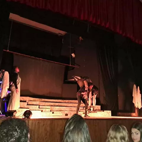 Representación teatral en interior con actores sobre el escenario y público observando.