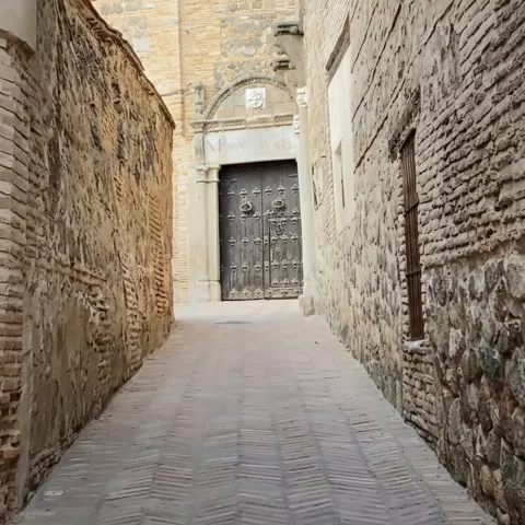 Callejón empedrado entre muros de piedra que conduce a una gran puerta.