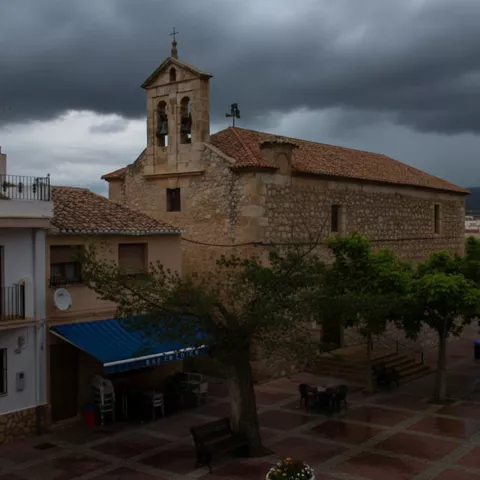 Iglesia de piedra en la plaza de La Recueja, con bancos y árboles en un ambiente tranquilo de pueblo.
