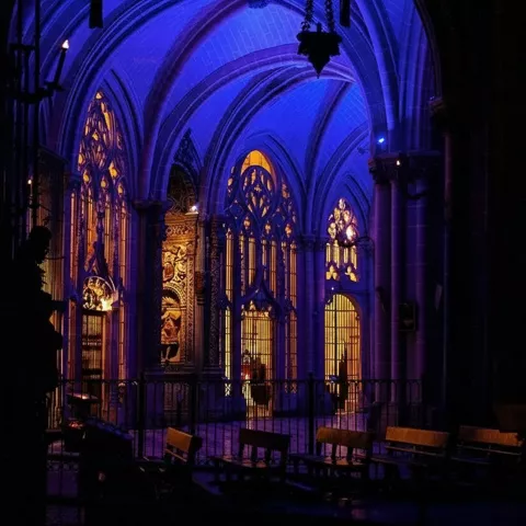 Nave gótica iluminada en azul y dorado con vitrales y bancos.