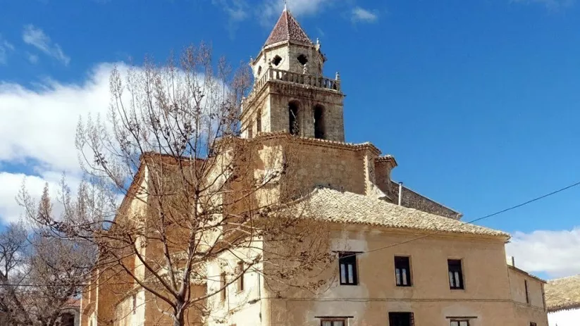 Iglesia de Mahora con torre campanario y fachada de piedra en un entorno urbano tranquilo.