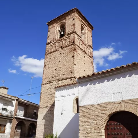 Primer plano de una torre de iglesia construida en ladrillo visto con un campanario, situada junto a un edificio blanco de baja altura.