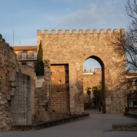 Entrada fortificada con arco y almenas, junto a restos de muros en una plaza.