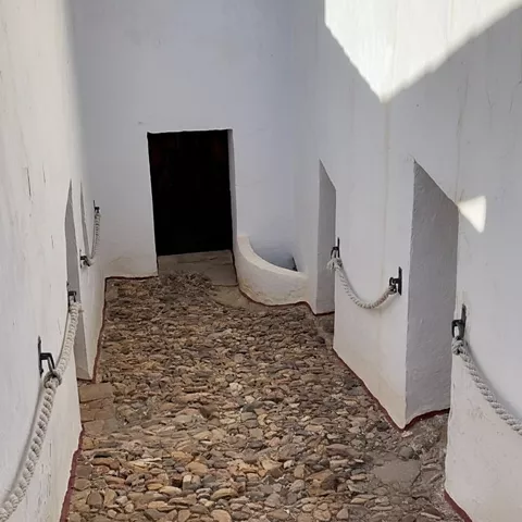 Pasadizo blanco con suelo de piedras y cuerdas a modo de pasamanos.