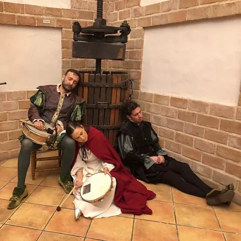 Personas con vestimenta de época sentadas junto a una prensa antigua en sala de ladrillo.