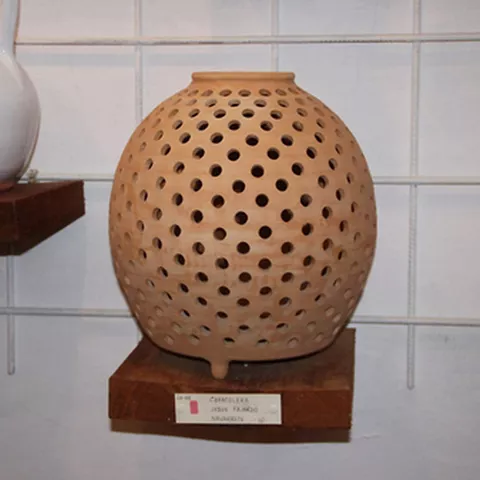 Vasija de cerámica perforada expuesta sobre una peana de madera.