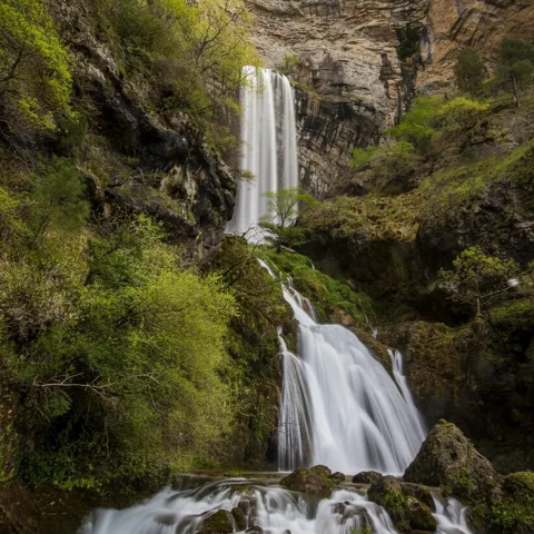 Cascada alta en un barranco rocoso, con vegetación densa y poza en la base.